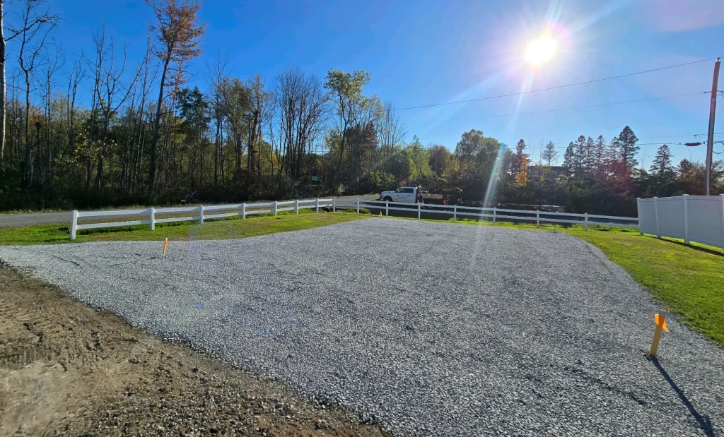 An empty gravel lot with a white fence and a forest in the background under a sunny blue sky.